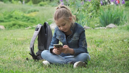 A pensive girl of nine years old, a schoolgirl, sits on the grass in the park. She holds a...