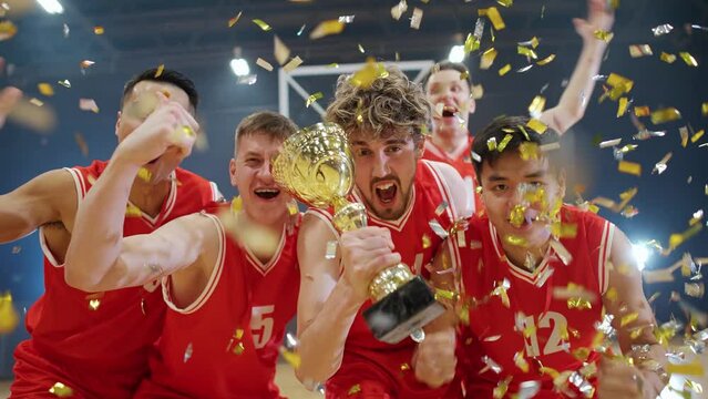 Slow motion, the red team of basketball players grejoices at the victory in the international championship, confetti is flying in the air, man holds the gold cup in her hands, emotions of victory.