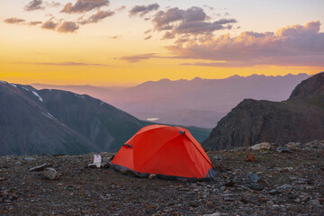 Scenic alpine landscape with tent at very high altitude with view to large mountains in orange dawn sky. Vivid orange tent with awesome view to high mountain range under cloudy sky in sunset colors.