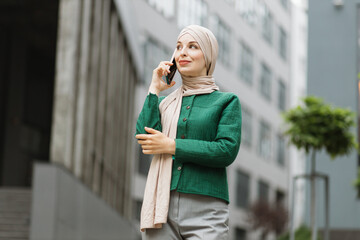 Fototapeta premium Muslim business woman in green jacket and hijab standing on street and talking on mobile. Confident female entrepreneur having working conversation on phone, outdoor on background of modern buildings