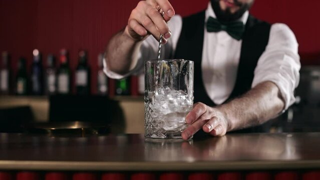 Close up of professional bartender stirring with long spoon ice cubes and alcoholic drink behind bar counter. Man in uniform preparing fresh cocktail at luxury restaurant.