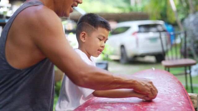 Happy Asian family grandfather teaching grandchild boy waxing surfboard surface. Senior man and little boy enjoy outdoor activity lifestyle water sport surfing together at the sea on summer vacation.