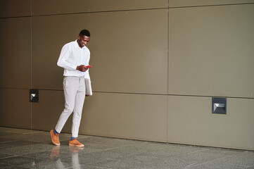 Full body of content African male entrepreneur in formal clothes texting message on cellphone while walking along wall in office building