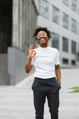 Beautiful young african american woman with black curly hairstyle. Beautiful girl in casual clothes in the street. Happy female wearing white t-shirt and dark pants showing sign ok.