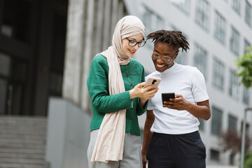 Charming woman in hijab withsmartphone and african female with phone standing together near office building. Two women colleagues using modern gadgets for work outdoors. - obrazy, fototapety, plakaty