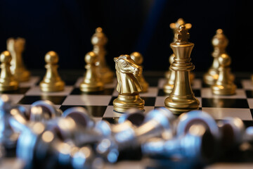Golden knight placed against many silver chess pieces on chessboard with businessman in background