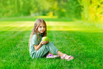 Fototapeta premium a happy child girl in the summer on the lawn with a green apple sits on the grass and smiles