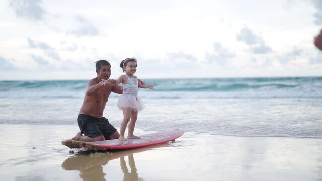4K Happy Asian family grandfather teaching little grandchild girl surfing on surfboard at the beach. Senior man and little girl enjoy outdoor activity lifestyle water sport surfing on summer vacation