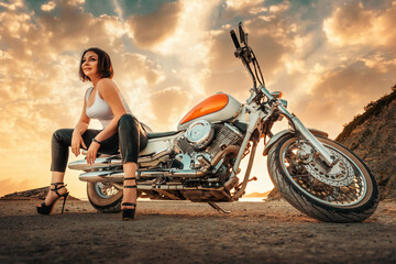 Plakat Beautiful sexy adult woman with a high heels and leather pants, posing sitting on motorcycle. Epic sunset sky on the background. Bottom view. The concept of Motorcyclist Day