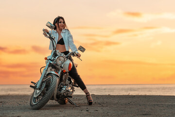 Fototapeta na wymiar Attractive sexy adult woman with a high heels and leather pants, posing confidently with motorcycle. Sunset sky on the background. Copy space. The concept of Motorcyclist Day