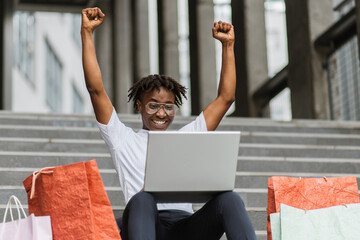 Pretty African American young woman, making online purchases using laptop sitting on the steps of...