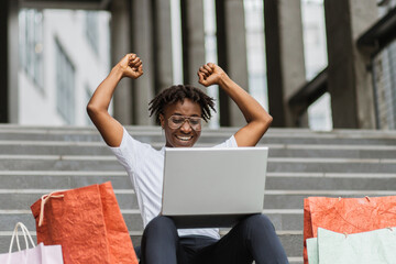 Pretty African American young woman, making online purchases using laptop sitting on the steps of...