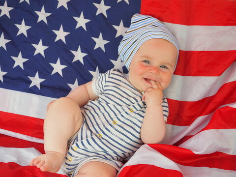 Cute Baby Is Lying On The American Flag. Happy Infant Plays Swinging On A Swing Against The Background Of The National Flag Of The United States Of America.