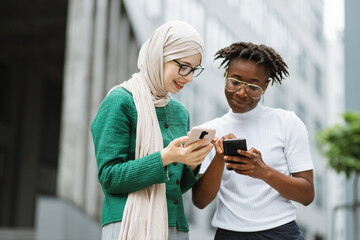 Charming woman in hijab withsmartphone and african female with phone standing together near office building. Two women colleagues using modern gadgets for work outdoors.