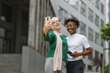 Two elegant women, one caucasian in muslim clothes and african american, friends smiling while looking at camera of mobile phone, making selfie portrait outdoors on urban background.