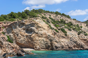 Fototapeta na wymiar Part of steep coast, layers of centuries-old rock are clearly visible, covered with green bushes and moss, clear sea, Ibiza, Balearic Islands, Spain