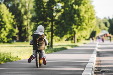Funny little boy riding a bike (running bike) outdoors. A happy child walks in the spring park. The kid is dressed in a fashionable vest and a tracksuit.
