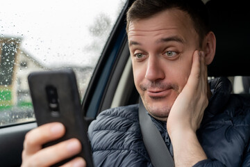 Millennial man shocked when see smartphone while sitting in car. Surprised businessman look at...