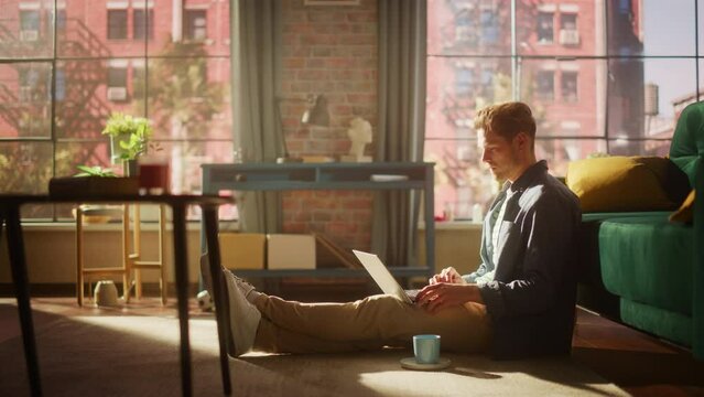 Portrait of a Handsome Young Adult Male Sitting on a Floor and Using Laptop Computer in Sunny Loft Living Room. Successful Man Working from Home in Bright Apartment.