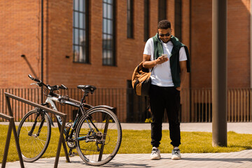 Trendy african american guy in sunglasses using smartphone while walking down the street in urban city, full length shot