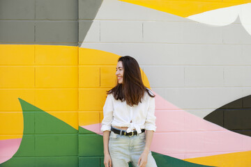 Young beautiful millennial woman standing by bright colorful green, yellow, pink wall, smiling and...