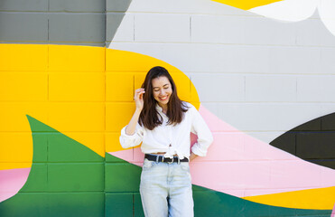 Young beautiful millennial woman standing by bright colorful green, yellow, pink wall, smiling and laughing - candid lifestyle ordinary moments