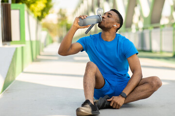 Handsome African American guy in sportswear drinks a bottle of water or a sports drink, sits on the...