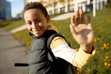 Side view of 8-years-old african american boy going to school, dressed in yellow hoodie and black puffer vest, carrying backpack, doing bye with hand, over background with school and green grass © Anatoliy Karlyuk