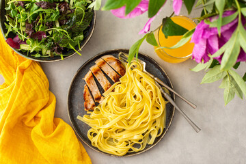 Hearty delicious lunch, pasta with fried chicken breast and fresh salad and orange juice, top view of the table with food and a vase of flowers