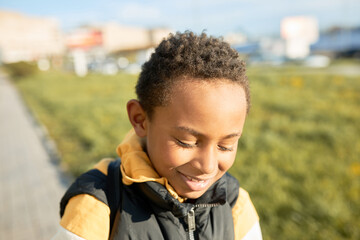 Horizontal outdoor portrait of shy african kid in hoodie and puffer vest looking down posing over cityscape with green lawn and sidewalk, walking in sunny evening. Happy carefree childhood