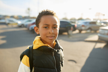 Portrait of handsome serious boy of african ethnicity in stylish outfit posing over parked cars, walking in city center, looking at camera, squinting in the sun. Urban lifestyle. Happy childhood - Powered by Adobe