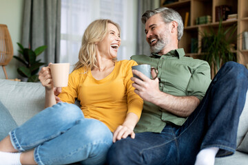 Joyful Middle Aged Spouses Relaxing On Couch With Coffee And Chatting