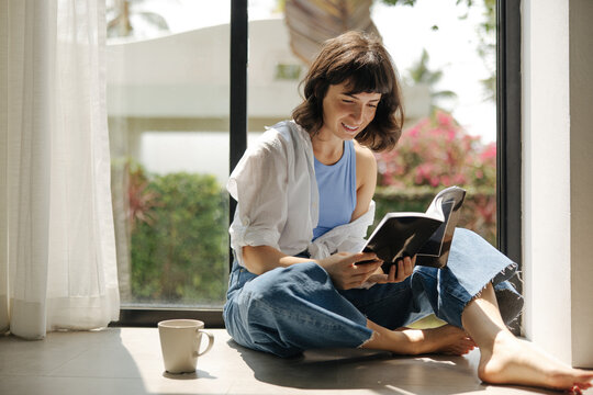 Attractive young caucasian brunette girl holding book sitting by window on warm day. Female student wearing casual clothes spend leisure time. Lifestyle relax education concept