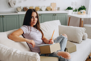 Obraz premium Positive Girl holding box and writing down notes, wearing white t-shirt, sitting on sofa in room
