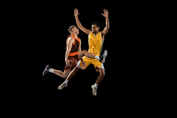 Dynamic portrait of two young men, professional basketball players in a jump, throwing ball into basket isolated over black studio background.