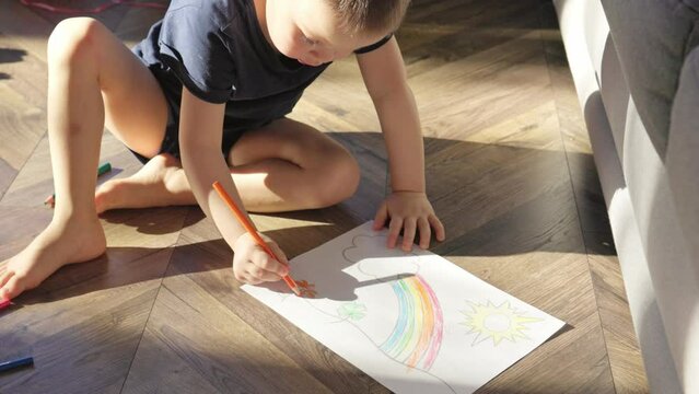 caucasian boy coloring rainbow drawing on wooden floor in living-room sunny day. little baby child sitting lying on floor at home, drawing picture with colorful pencils crayons. concept happy family