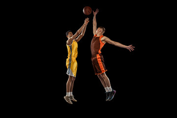Fototapeta na wymiar Dynamic portrait of two young men, professional basketball players in a jump, throwing ball into basket isolated over black studio background.