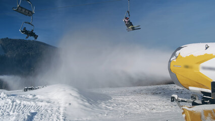 Snow cannon makes artificial snow on background of working ski lift with people, going to mountain...