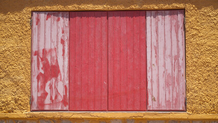 Ventana cerrada de tablones de madera pintados de rojo en pared amarilla de casa de playa