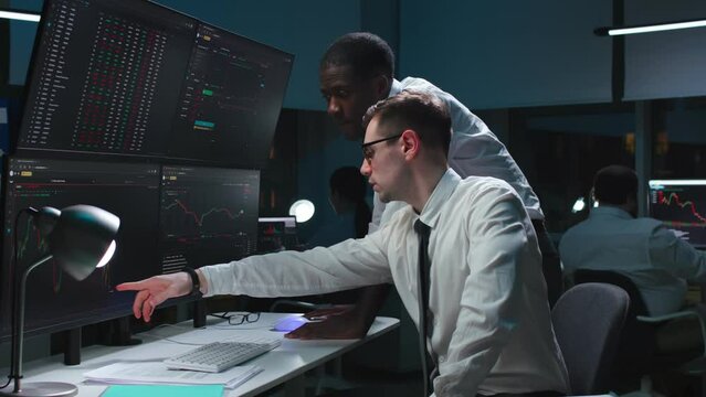 African-American and caucasian brokers analyze real-time stock market graphs on multiple monitors