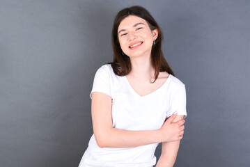 Fototapeta premium People, lifestyle, youth and happiness concept. Shy pretty young beautiful Caucasian woman wearing white T-shirt over studio grey wall, feeling happy hugging herself.