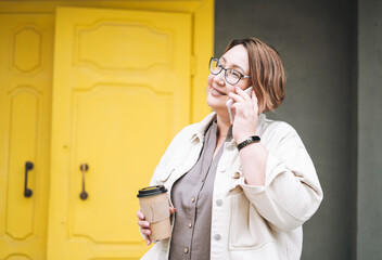 Adult brunette middle aged woman plus size body positive in casual clothes with paper cup of coffee using mobile phone near yellow door at city