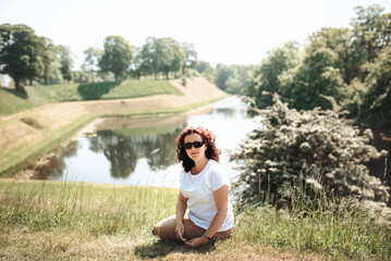 Girl-traveler sits on green grass and looks straight. Woman enjoys nature and sits on the banks of moat around Castellet Castle. XVII century fortress with museum in Copenhagen, Denmark. Soft focus