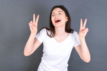 Isolated shot of cheerful young beautiful Caucasian woman wearing white T-shirt over studio grey wall makes peace or victory sign with both hands, feels cool.