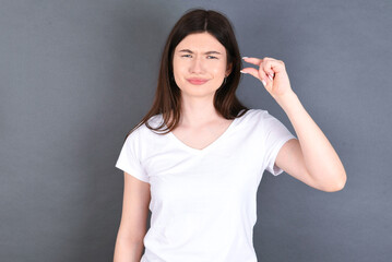 Upset young beautiful Caucasian woman wearing white T-shirt over studio grey wall shapes little...