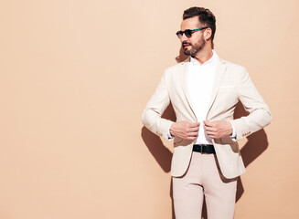 Portrait of handsome smiling stylish hipster lambersexual model. Sexy modern man dressed in elegant white suit. Fashion male with curly hairstyle posing in studio near beige wall. In sunglasses