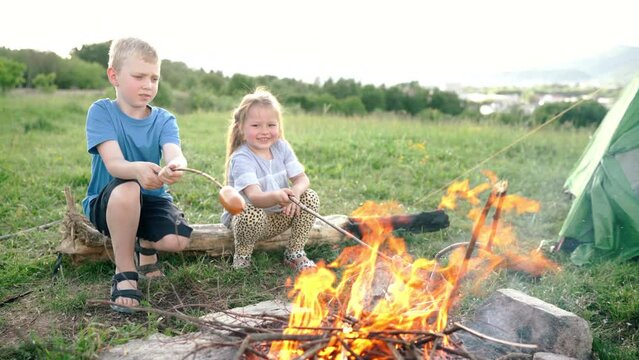 Brother and sister have conversation and cheerfully smiling while they roasting a  sausages on the sticks over the campfire flame in tent camp. Happy family or outdoor picnic activities concept image.