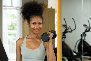 Fototapeta na wymiar Young curly black hair woman enjoy holiday in fitness center. Lifting barbell with one hand while smiling.