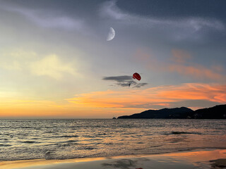 Amazing colorful sunset. Cape in the distance. Tropical beach. Parachute in the sky above the sea. Water sport activity. Parasailing. Clouds, crescent and stars on the skies. Reflection of sun glow. 