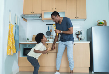 Funny portrait of smiling African American father and his son singing songs, dancing to music while cooking food together, using whisk and spatula as microphone in kitchen at home.
